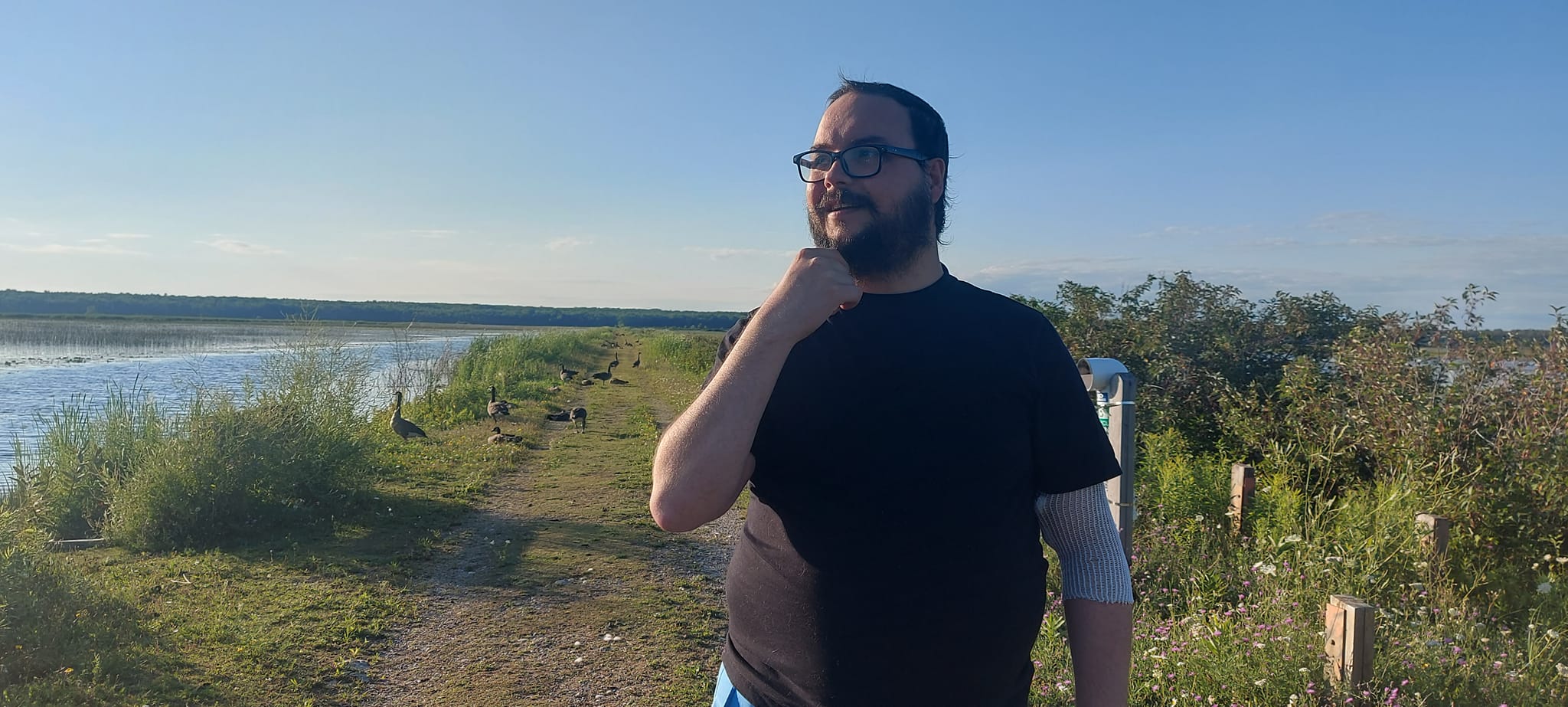 A man short hair and bushy facial hair stands on a dyke in a marsh in front of a dozen Canada geese